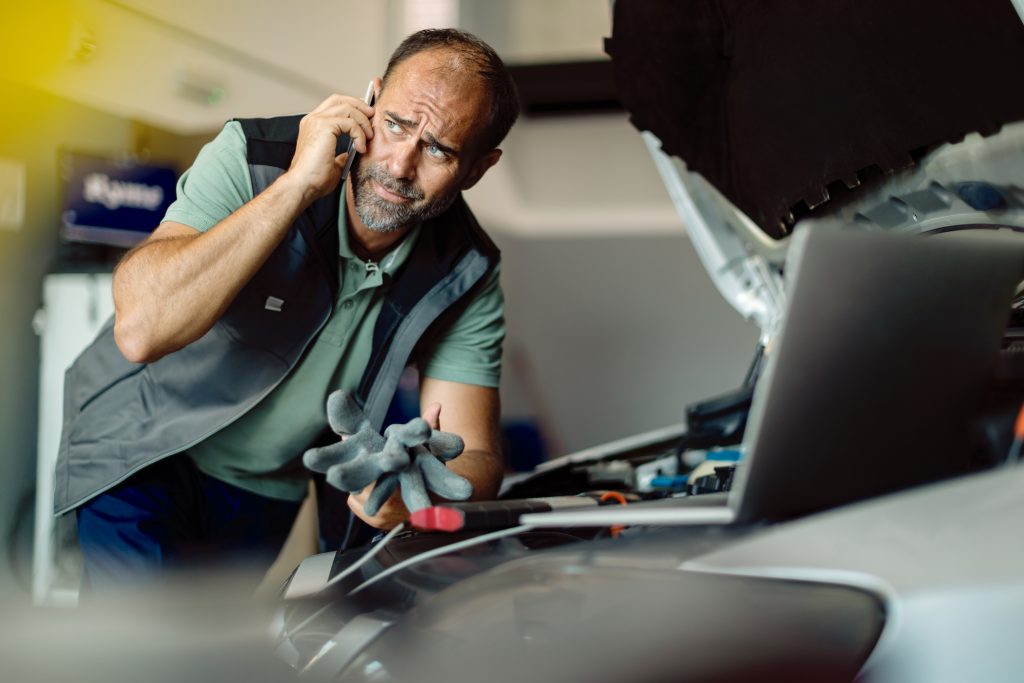 Mid adult car mechanic communicating on mobile phone while repairing car in a workshop.